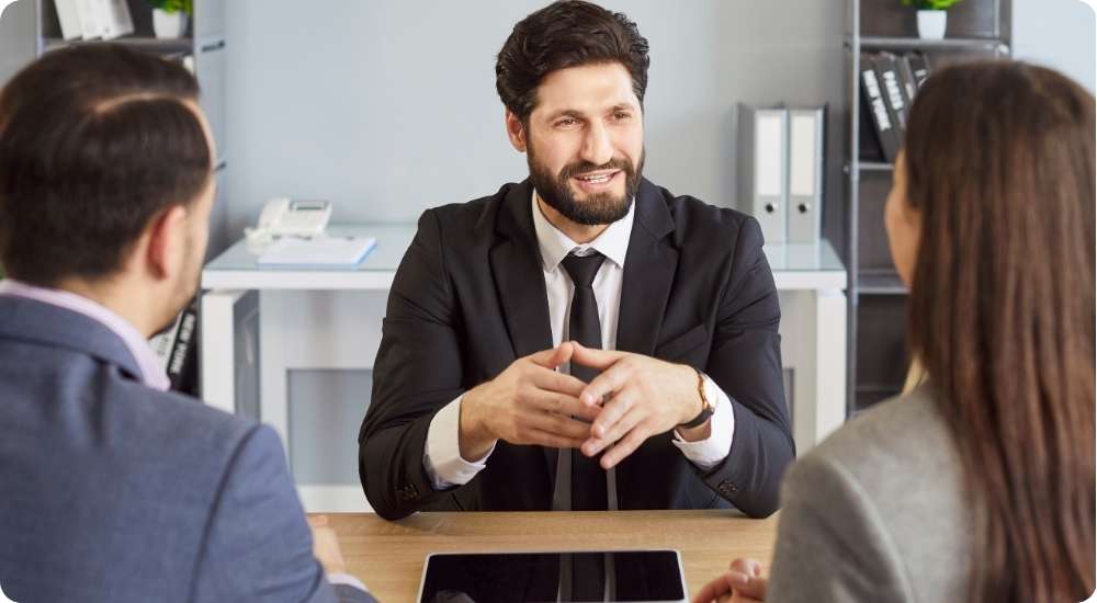 Three professionals sit around a wooden office table; the man in the center wears a suit and actively speaks while the two individuals facing away listen, symbolizing a panel job interview for a senior role.