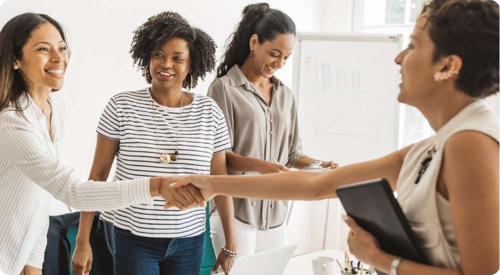 A diverse group of four professional women in a brightly lit office environment, with two of them shaking hands and smiling, symbolizing a successful new hire, interview, or business agreement.