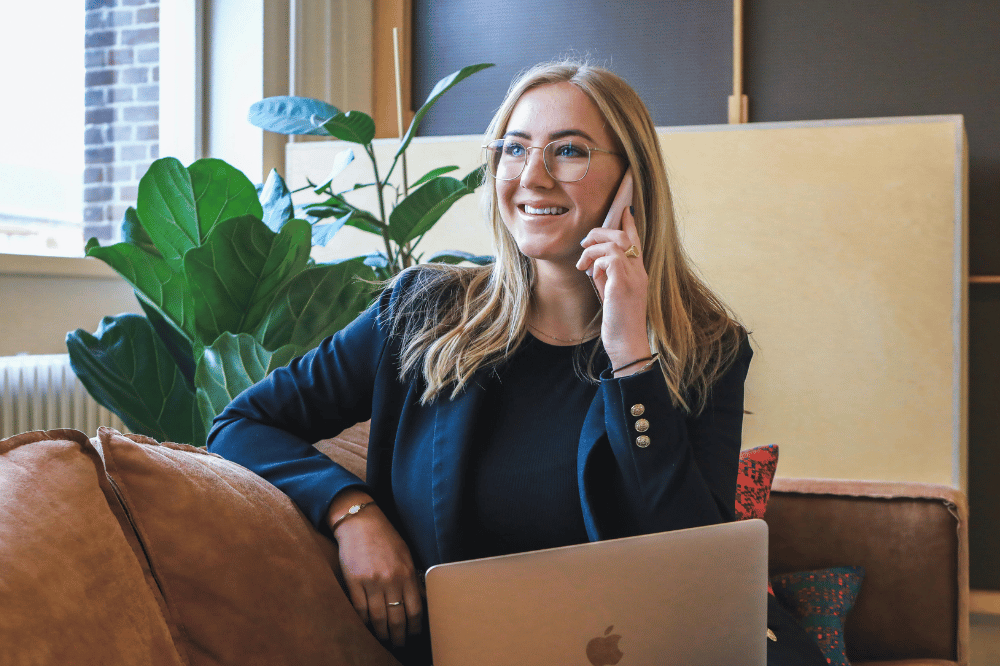 A smiling woman with blonde hair and glasses, wearing a navy blazer, sits on a leather couch while talking on a smartphone with an open laptop in front of her.