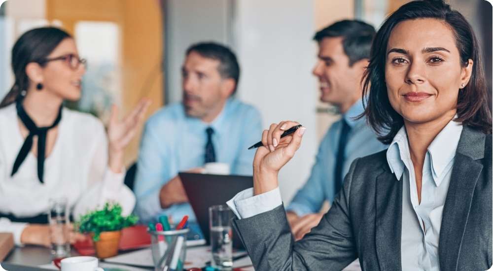 A female business executive in a grey suit looks confidently at the camera while three other diverse professionals engage in a meeting and discussion around a boardroom table, symbolizing leadership development and teamwork.