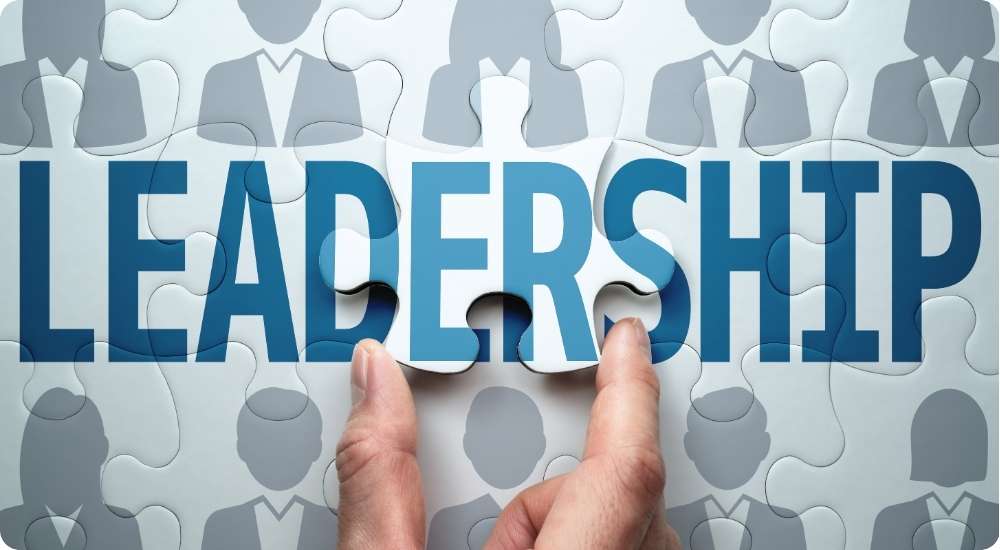 A close up photograph of a person's hands placing the final puzzle piece into the word "LEADERSHIP," which is set against a background of grey human silhouette icons, symbolizing the building of a leadership pipeline.