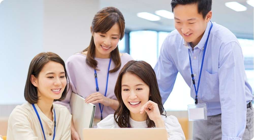 A diverse group of four professionals in a modern, brightly lit office environment leaning in together, smiling, and happily collaborating around a laptop computer.