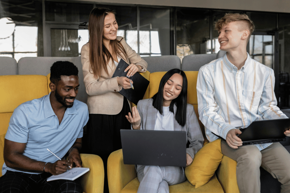 A group of four diverse young professionals smile and chat while collaborating in a modern lounge area with yellow seating, using laptops and a notebook.
