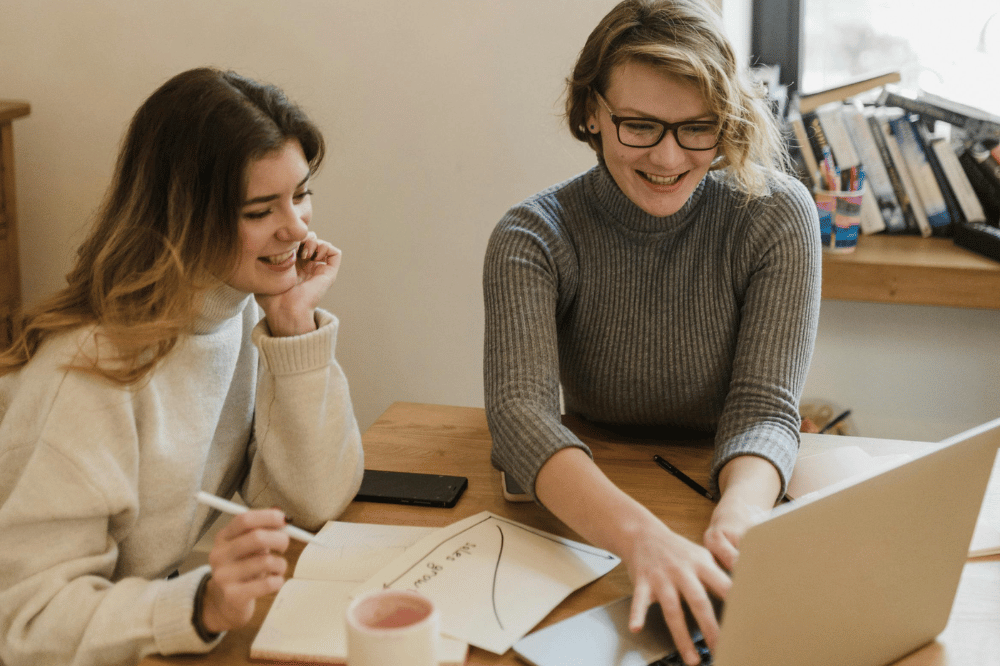 Two smiling women sit at a wooden desk with a laptop and notebook, collaborating and pointing at the screen in a bright room with bookshelves.