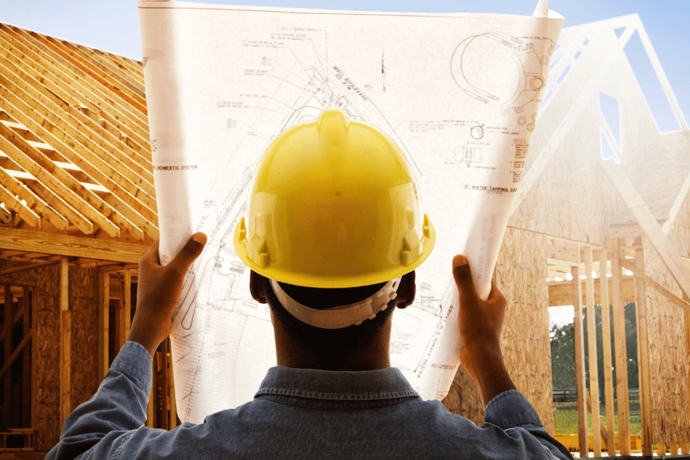 Back view of a construction worker wearing a yellow hard hat, holding up and reviewing architectural blueprints against the backdrop of a house's wooden frame under construction.