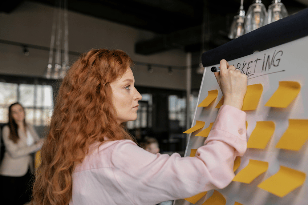 A woman with long, wavy red hair writes "MARKETING" on a whiteboard covered in yellow sticky notes during a brainstorming session in an office.