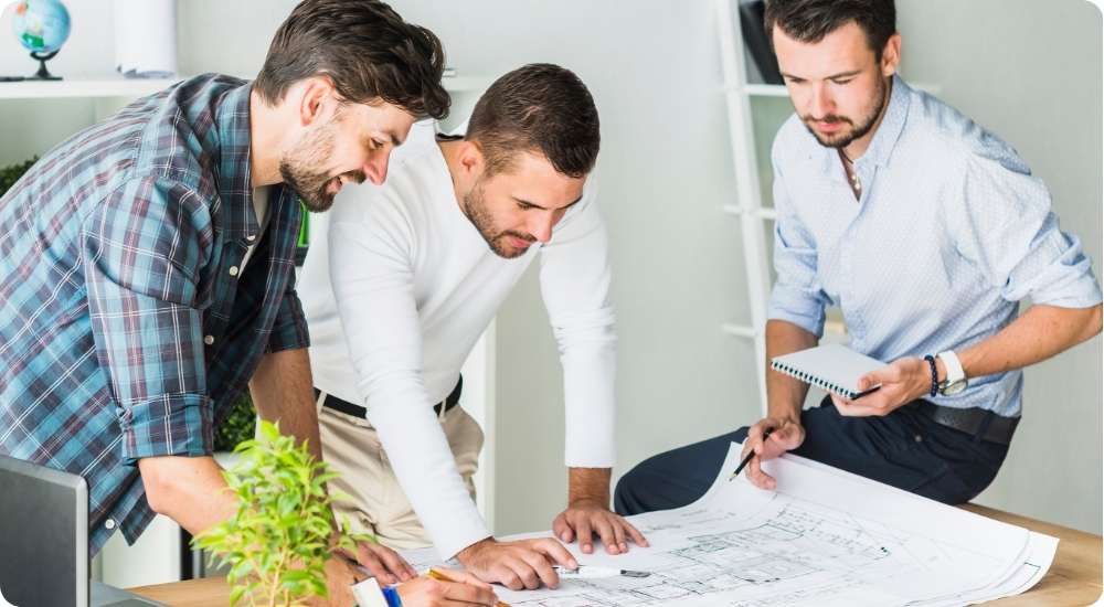 Three male professionals, likely architects or engineers, review detailed blueprints spread across a table in an office, collaborating on a design project.
