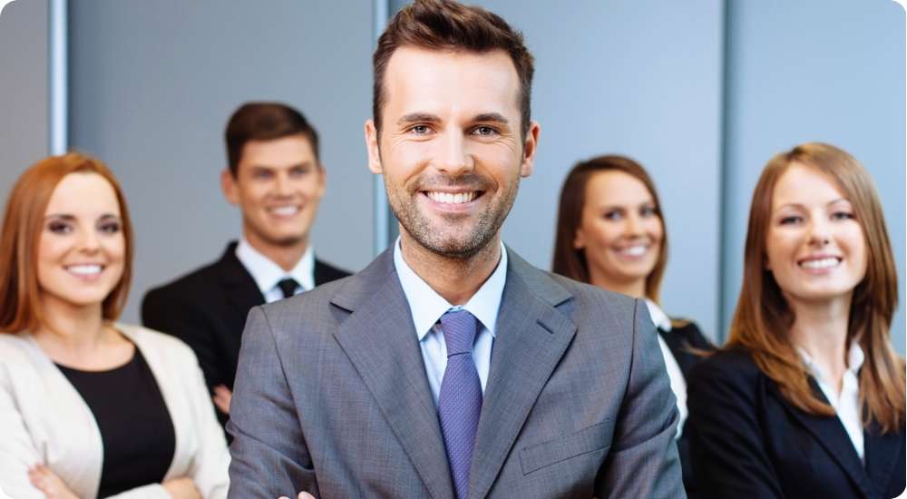 A diverse group of five smiling business professionals in formal attire standing together in an office setting, with a man in a grey suit prominently featured in the foreground.