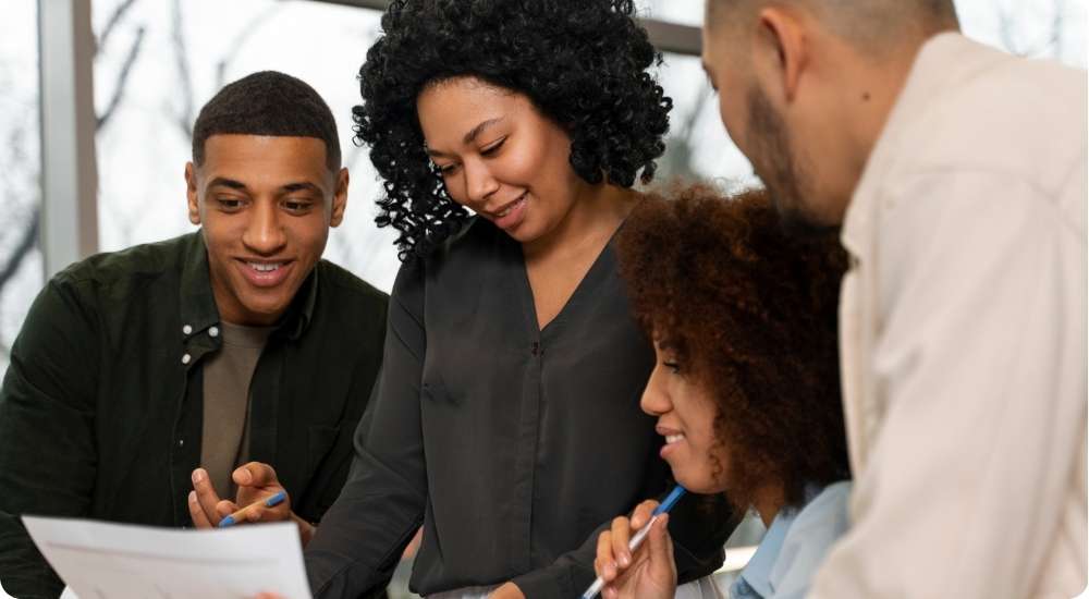 A diverse group of four business professionals in a casual meeting setting, collaborating and reviewing paperwork on a table, symbolizing teamwork and a positive work environment.
