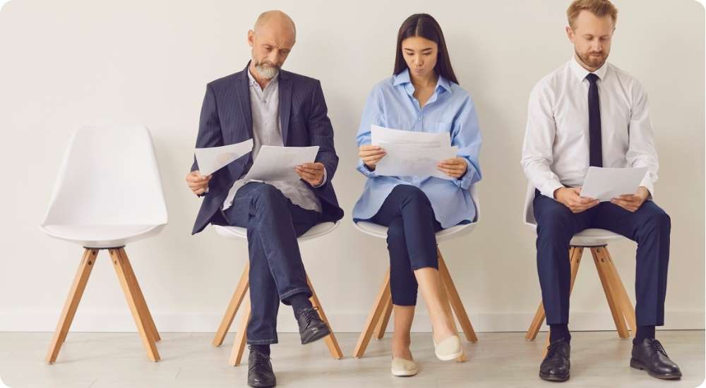 Three diverse business professionals, two men and one woman, sit on modern white chairs in a waiting room, reviewing documents before an interview or meeting, symbolizing the executive hiring process.