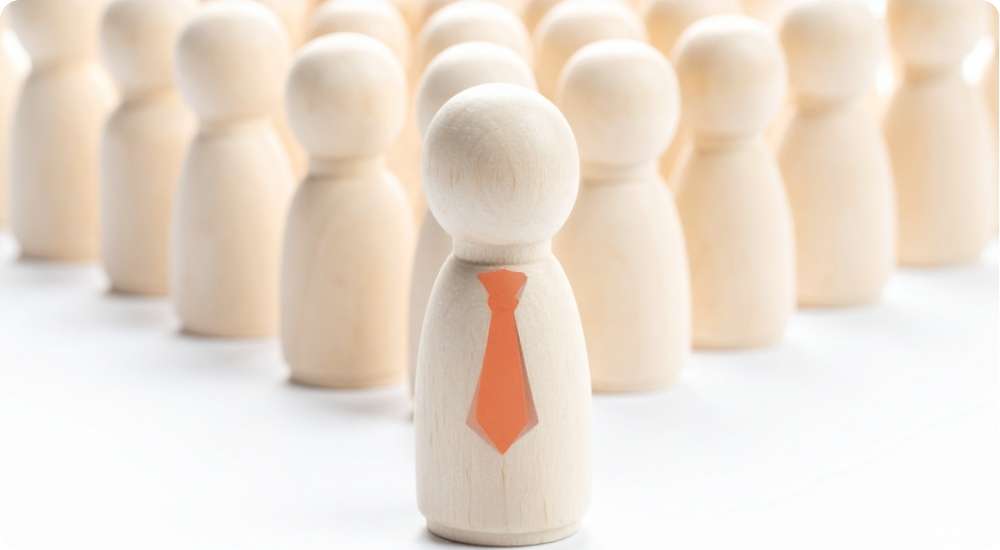 A close-up of a wooden figure wearing an orange tie, standing in the foreground, with many other blurred, identical wooden figures behind it, symbolizing leadership, key personnel selection, and overall workforce management.
