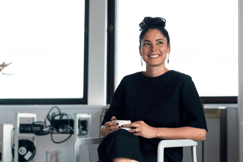 A woman with dark hair tied in a bun and wearing long earrings and a black shirt sits in a modern office, smiling as she looks off-camera while holding a white cup.