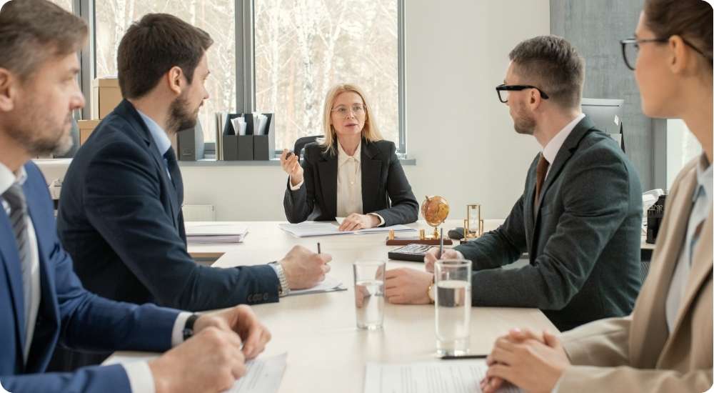 A group of diverse professionals in formal business attire sitting around a conference table during a meeting, representing a team collaborating on hiring roadmaps and strategic workforce planning for a modern tech firm.