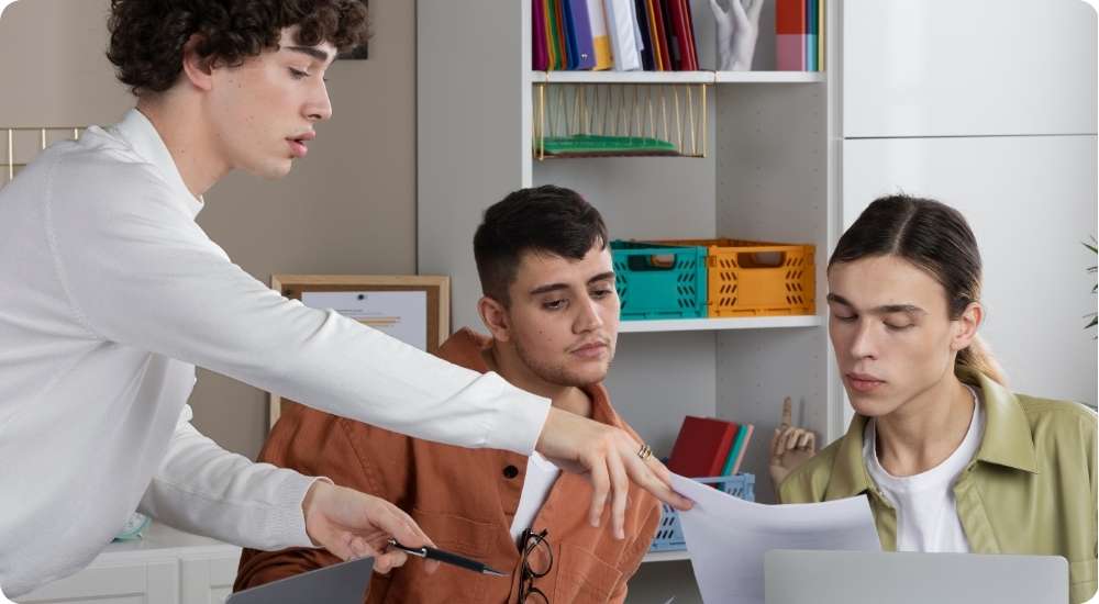 Three young professionals are collaborating around a desk in a modern office. The person on the left is pointing with a pen at a document and explaining content to two colleagues, symbolizing teamwork, mentorship, and a learning environment.