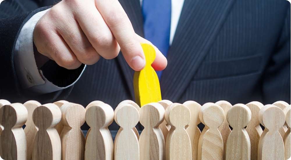 A close-up of a hand in a suit reaching down to select a yellow wooden figure from a crowd of many identical, light-wood figures, symbolizing the hiring process, human resource selection, and finding the right person for a job in a skills-based economy.