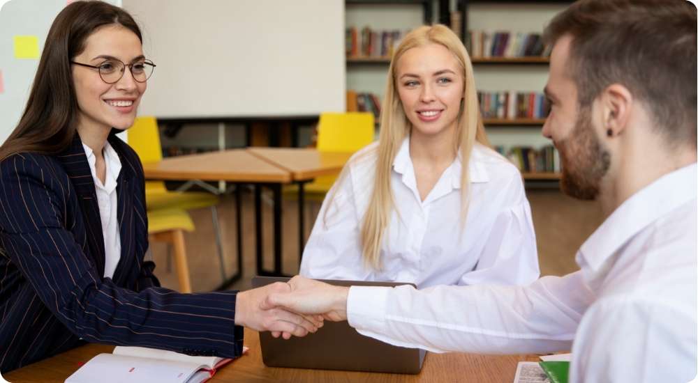 A close-up shot of a male and female business professional shaking hands across a conference table with open notebooks, symbolizing a successful partnership, a signed agreement, or a new employee being welcomed to a leadership team.
