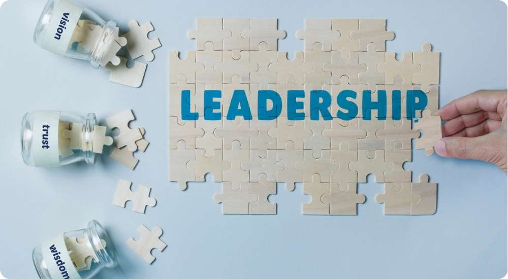 A hand places the final puzzle piece to complete the word LEADERSHIP. Three glass jars nearby contain puzzle pieces labeled vision, trust, and wisdom, illustrating the core components of effective leadership skills.