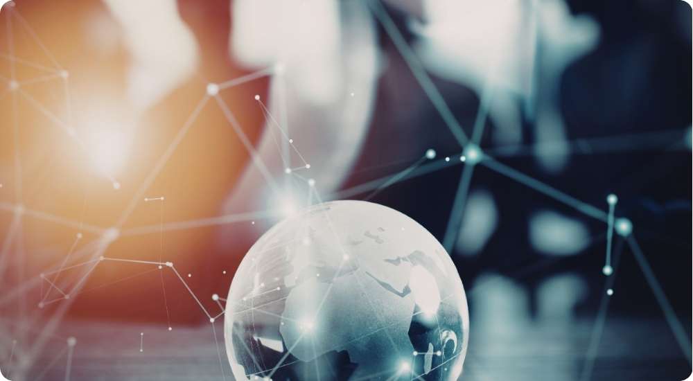 A close-up shot of a glass globe with a glowing digital network overlay sits on a desk. In the blurred background, the silhouettes of business professionals are visible, symbolizing global business connectivity and technological collaboration.