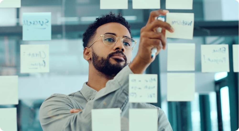 A close-up photograph of a thoughtful man in glasses organizing white and blue sticky notes on a glass wall, symbolizing strategic planning and long-term business lessons.