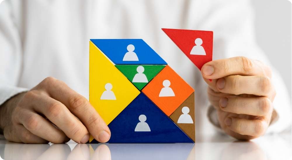 A close-up photograph of a person's hands placing a final red triangular puzzle piece onto a multi-colored tangram puzzle featuring white human icons, symbolizing the assembly or management of a diverse team structure.