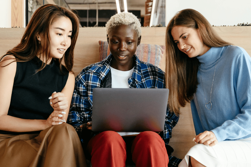 Three diverse women sit closely together, smiling as they look at and discuss something on a laptop screen.