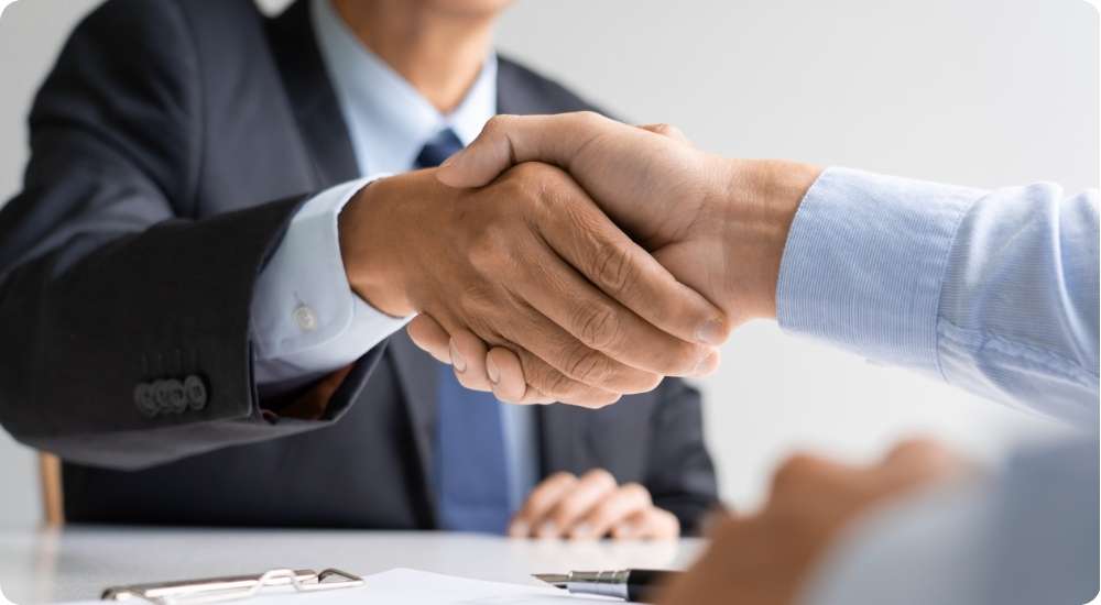 A close-up photograph of two business professionals in formal attire shaking hands over a meeting table, symbolizing agreement, partnership, an interview conclusion, or a successful new hire.