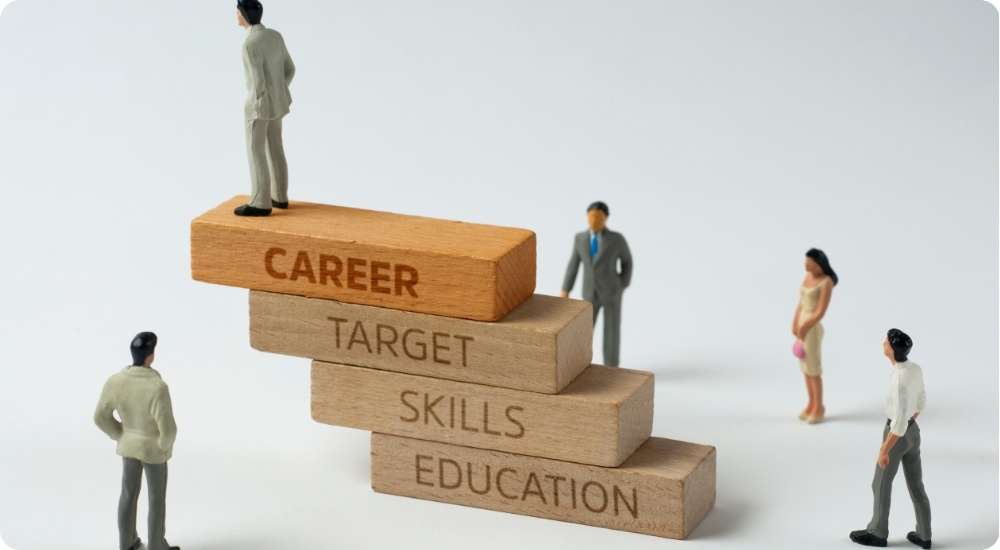 A close-up of a human figure standing at the top of a four-step wooden block pyramid. The blocks are labeled 'EDUCATION', 'SKILLS', 'TARGET', and 'CAREER', illustrating the pathway and growth toward career success.