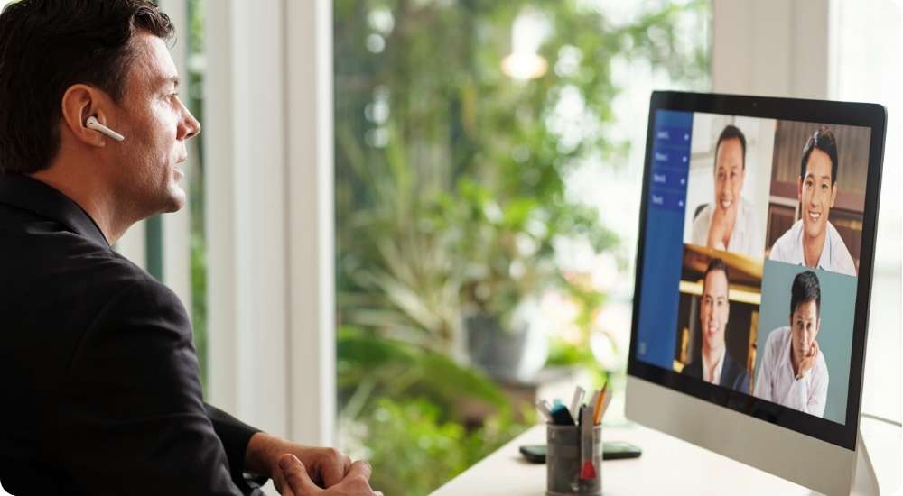 A professional man wearing a wireless earbud sits at a desk and participates in a multi-person video conference call on a computer monitor, symbolizing remote leadership skills.