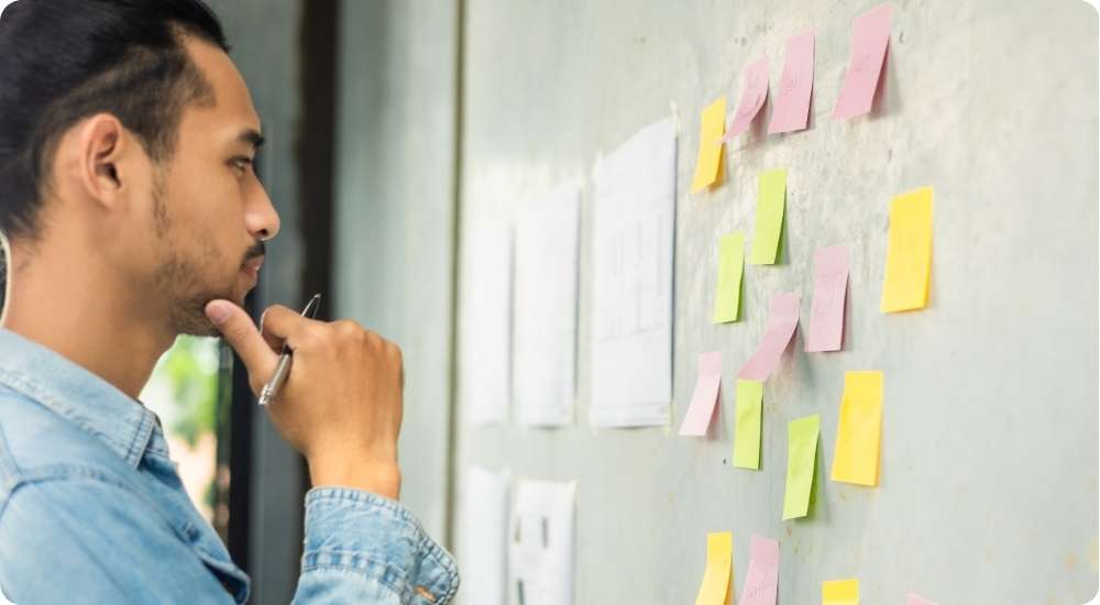 A close-up photograph of a man thoughtfully analyzing a grey wall covered with colorful sticky notes, representing a brainstorming session or strategic planning process.