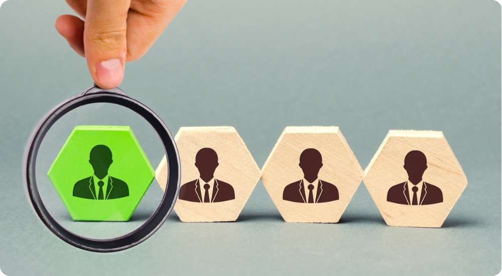 Magnifying glass highlights a green hexagon with a person icon from a line of grey hexagon employee blocks.