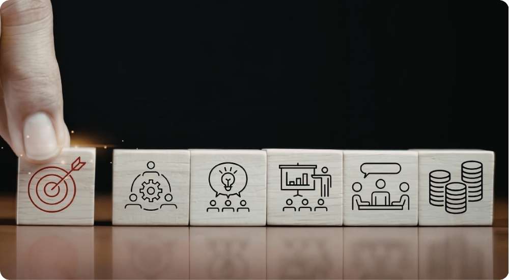 A close-up view of a hand placing a wooden block with a target icon in a sequence of blocks with icons for teamwork, ideas, presentation, meeting, and finance, symbolizing strategic business and hiring objectives.