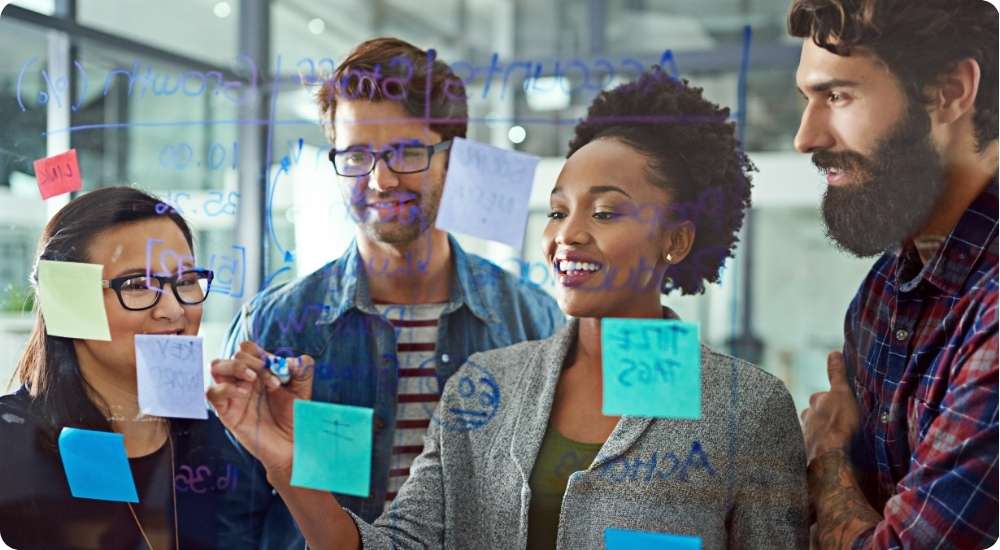 A diverse team of four business professionals collaboratively adding colorful sticky notes and writing on a glass whiteboard during a strategic planning meeting.
