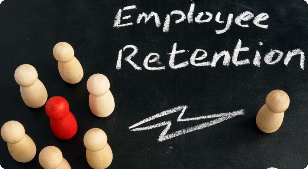 A red wooden figure stands out in a group of several plain wooden figures on a black background, with the words Employee Retention written in chalk. A chalk lightning bolt is drawn near the red figure, symbolizing standing out, key talent, or employee turnover.