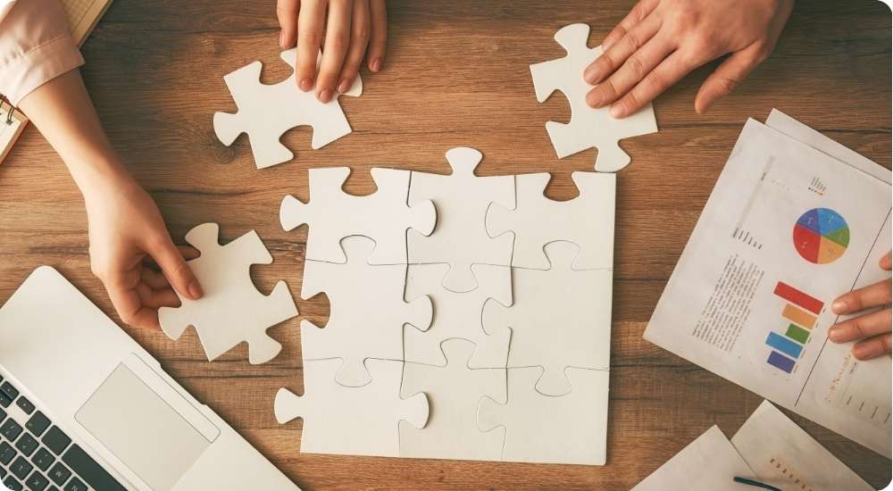 An overhead shot of several people collaborating to assemble a large white jigsaw puzzle on a wooden table, with a laptop and business report nearby, symbolizing teamwork and strategic planning.