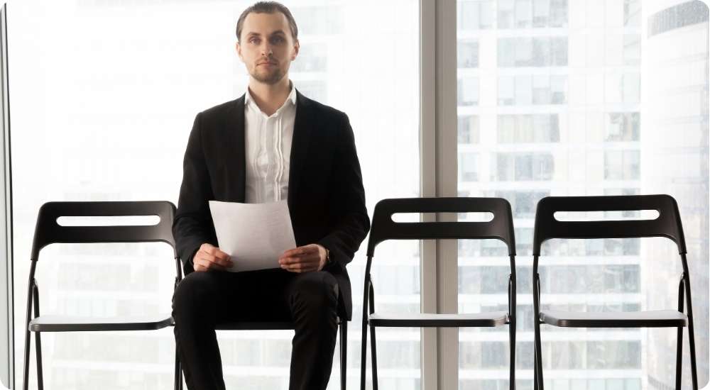 A male job candidate wearing a black suit and white shirt sits alone in a modern office waiting room holding a document, with several empty chairs beside him, illustrating the job application and interview process.