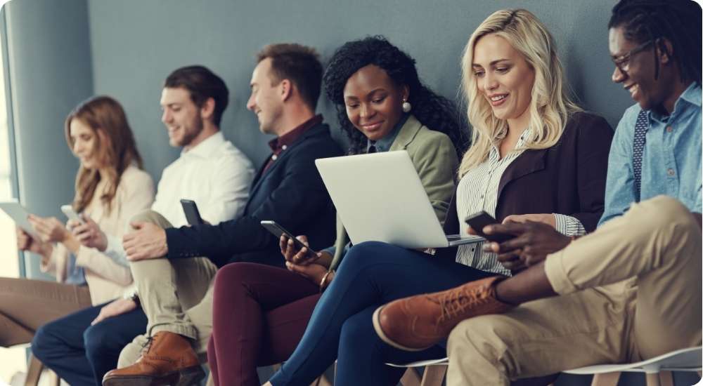 A diverse group of five professionals sitting in a modern office waiting area, with most individuals engaged with their smartphones or a laptop, appearing to be candidates waiting for an interview.
