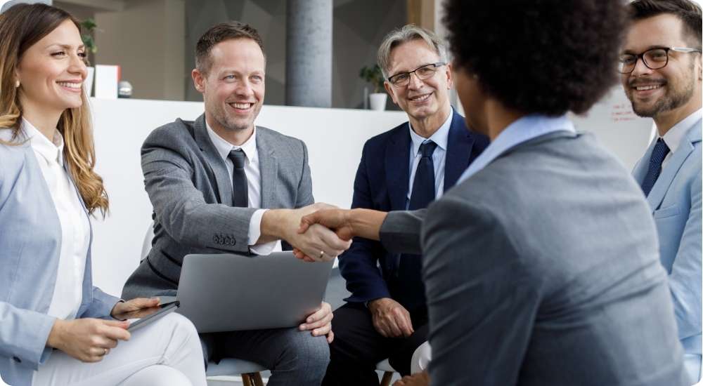 A diverse group of business professionals sitting around an office table, smiling as two individuals shake hands over a laptop, symbolizing a strategic partnership or agreement.