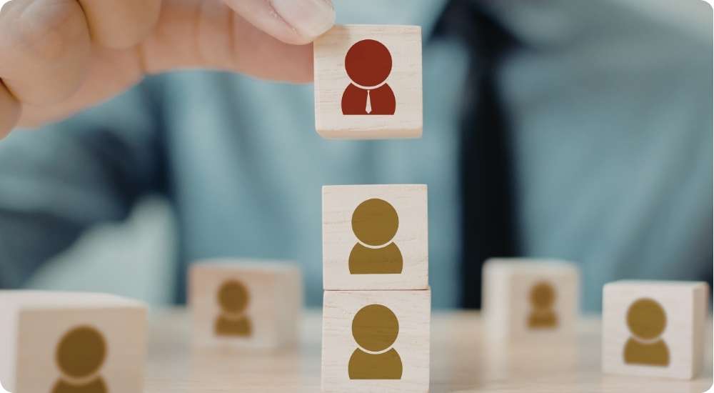A person's hand placing a small wooden block featuring a red human silhouette icon atop a stack of other blocks with grey icons, symbolizing team building, hierarchy, or the role of leadership in distributed teams.