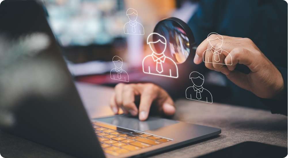 A business professional uses a laptop and holds a magnifying glass over digital outlines of potential employees, symbolizing the use of data and analysis to make informed human resource and workforce decisions.