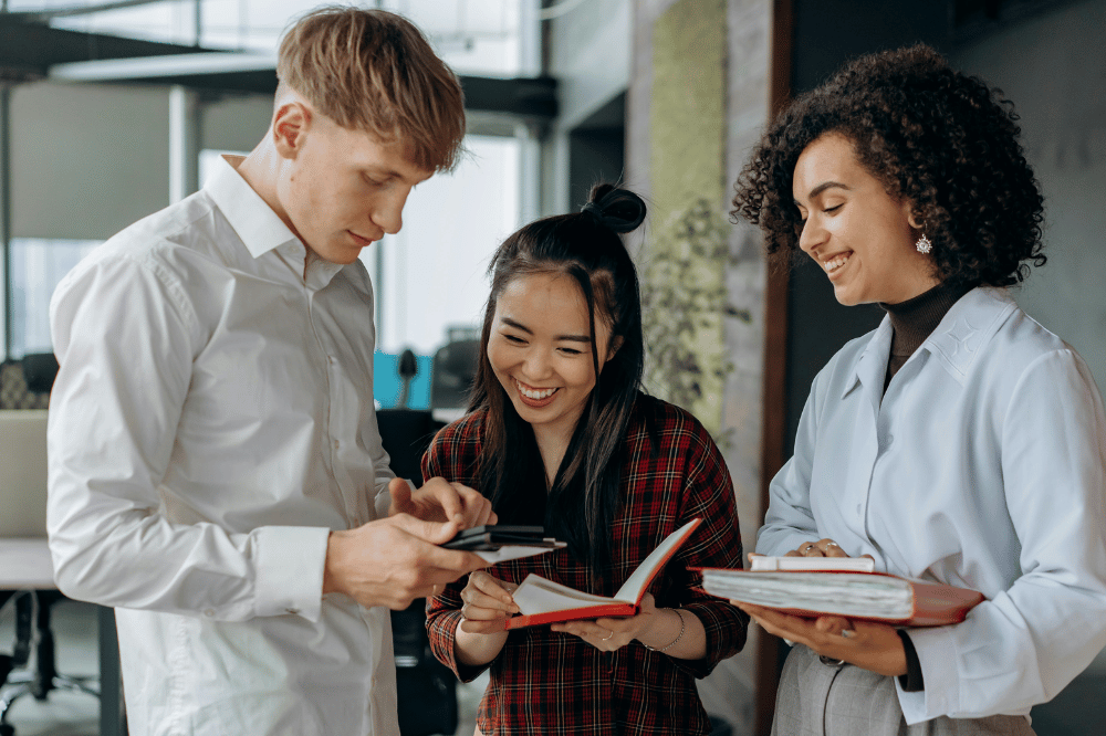 Three diverse colleagues stand together in a modern office, smiling as they collaborate and review information on a smartphone and in open notebooks.