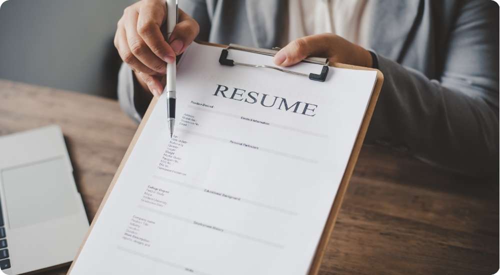 A person's hands holding a clipboard with a resume attached, while a pen points toward the document's content, symbolizing a job interview or the process of reviewing a candidate's qualifications.