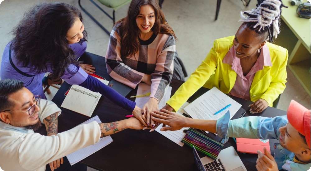 A diverse group of five smiling professionals in a modern office meeting, placing their hands in the center of the table in a gesture of unity, collaboration, and teamwork.