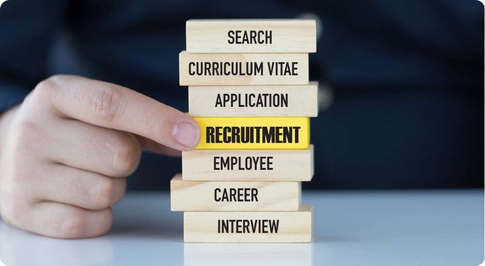 A person's hand touches a stack of wooden blocks with recruitment terms: search, curriculum vitae, application, recruitment, employee, career, interview. The "recruitment" block is highlighted in yellow.