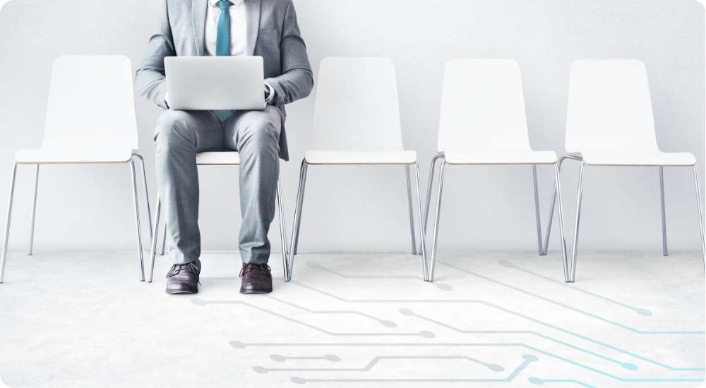 A businessman sits with a laptop in a waiting area with several empty chairs and a circuit board graphic on the floor, illustrating the human aspect of tech recruitment.