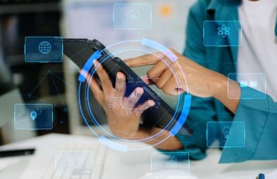 A person's hands using a tablet with a futuristic, data-oriented interface overlaid with icons like a globe, atom, and lightbulb, suggesting technology, data analysis, or AI.