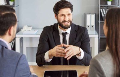 Three professionals sit around a wooden office table; the man in the center wears a suit and actively speaks while the two individuals facing away listen, symbolizing a panel job interview for a senior role.