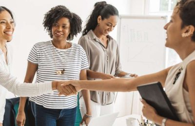 A diverse group of four professional women in a brightly lit office environment, with two of them shaking hands and smiling, symbolizing a successful new hire, interview, or business agreement.