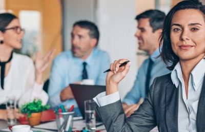 A female business executive in a grey suit looks confidently at the camera while three other diverse professionals engage in a meeting and discussion around a boardroom table, symbolizing leadership development and teamwork.