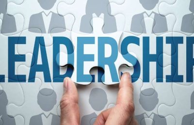 A close up photograph of a person's hands placing the final puzzle piece into the word "LEADERSHIP," which is set against a background of grey human silhouette icons, symbolizing the building of a leadership pipeline.