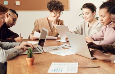 A diverse group of five business professionals works together around a conference table with laptops, papers, and a small cactus plant, illustrating team collaboration and a positive work environment.